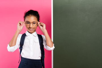 smiling african american schoolgirl with backpack near empty chalkboard on pink background