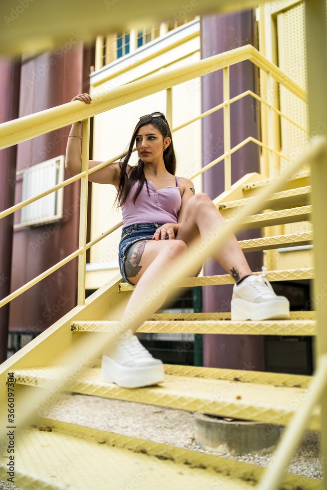 Poster Rebellious caucasian female with tattoos and in jeans skirt posing while sitting on the stairs