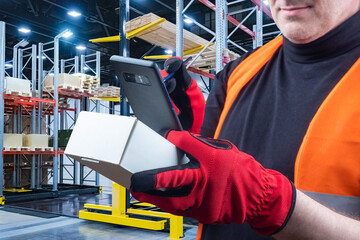 Fototapeta premium Man uses application for inventory control. Man in a warehouse holds phone in his hands. Storekeeper uses application on phone. Storekeeper photographs a small box. Application for warehouse business