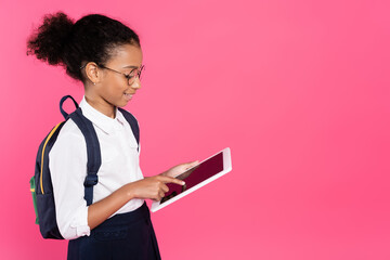 smiling african american schoolgirl in glasses with backpack using digital tablet isolated on pink