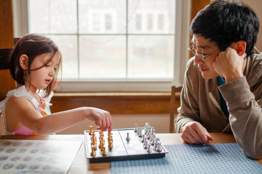 A small child plays chess with smiling father in window light