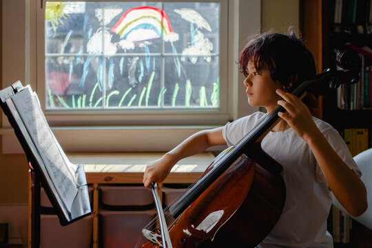 A focused boy sits in front of painted window practicing cello