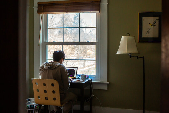 A man sits at desk in front of window in bedroom working on a computer