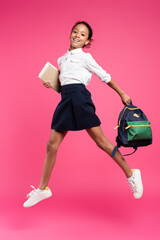 Fototapeta full length view of smiling african american schoolgirl jumping with backpack and books on pink background obraz
