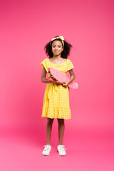 full length view of smiling curly african american child in yellow outfit with penny board on pink background