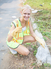 Happy female volunteer worker picking up plastic trash smiling and giving a thumbs up to the camera while wearing a reflective safety vest and gloves.