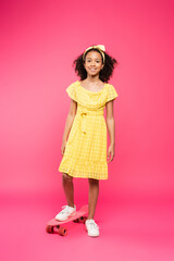 full length view of smiling curly african american child in yellow outfit on penny board on pink background