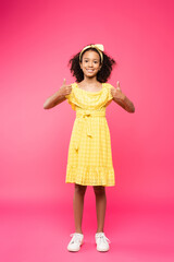 full length view of smiling curly african american child in yellow outfit showing thumbs up on pink background
