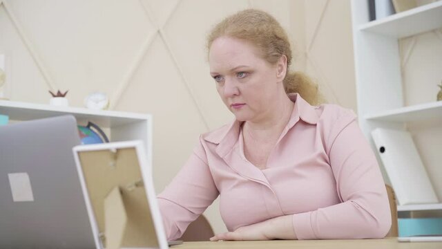Adult Caucasian woman typing on laptop keyboard and looking at picture in frame. Portrait of senior female employee working in office. Lifestyle, femininity.