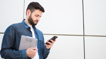 Serious brunette young man using mobile phone app texting outside of office in urban city on grey wall background.Focused caucasian businessman student in denim look at smartphone for business work