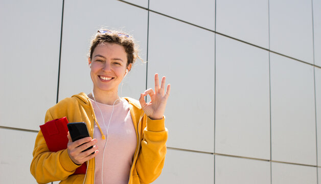 Smiling young woman in headphones showing ok okay gesture.Happy student hipster girl holding smartphone,listening audio course standing outdoor behind gray wall.Approval sign.Body language.Copy space