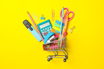 Shopping trolley with school supplies on yellow background, space for text