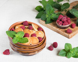 Trendy food pancake cereal, mini pancakes in a bowl with fresh raspberries and mint on a light textile background, selective focus
