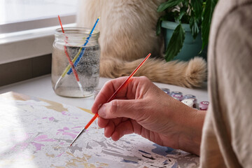 The older woman's hand holds a brush. She painting with acrylics by numbers on canvas.