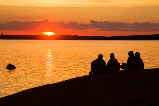 Four unknown human silhouettes are sitting on the shore and watching a beautiful sunset