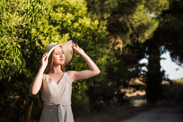 Young woman relaxing and enjoying the sun walking for the field. Young blonde woman with long hair dressed in a hat and striped dress
