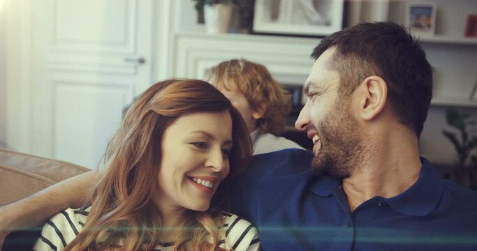 Young mom and dad are relaxing together sitting in living room on couch. Little son plays with parents hugging their necks.