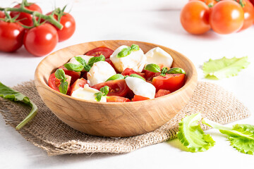 Caprese salad with fresh tomatoes, homemade mozzarella and basil in the wooden bowl on white marble table close up. 