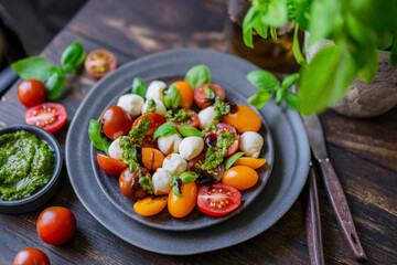 Caprese salad with fresh cherry tomatoes, mini mozzarella, pesto, basil, olive oil and balsamic sauce on blue plate, wooden rustic table. Mediterranean diet concept. Italian healthy food. Copy space.