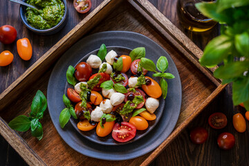 Caprese salad with fresh cherry tomatoes, mini mozzarella, pesto, basil, olive oil and balsamic sauce on blue plate, wooden rustic table. Mediterranean diet concept. Italian healthy food. Copy space.