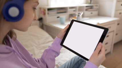 Close up teenage girl wearing headphones looking at white empty mockup tablet screen, watching video or webinar, playing mobile device game, chatting in social network online, making video call