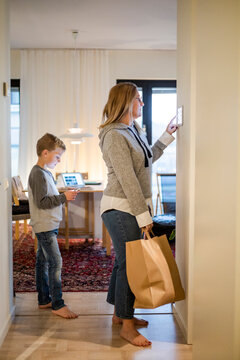 Mother with shopping bag using digital tablet on wall while son standing at home