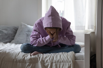 Unhappy depressed teenage girl wearing sweatshirt sitting on bed alone, feeling lonely and...