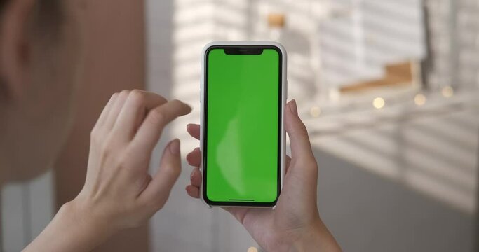 Girl using smartphone with green screen in living room watching movie, video content. Close up woman's hand holding smart phone in a vertical position. Indoors. Touch screen hand gestures. Closeup