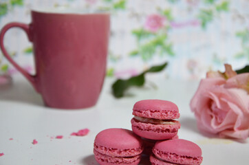 Three pink almond macaroons and mug with coffee on a white wooden background next to a pale pink rose. good morning concept
