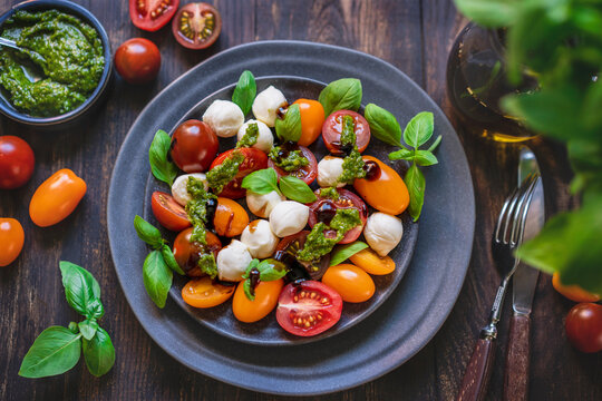 Caprese salad with fresh cherry tomatoes, mini mozzarella, pesto, basil, olive oil and balsamic sauce on blue plate, wooden rustic table. Mediterranean diet concept. Italian healthy food. Copy space.