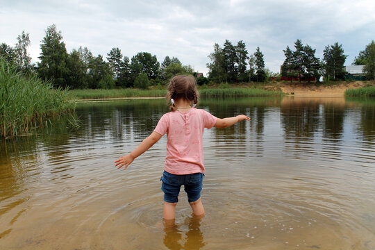 A Little Girl Plays Near The Lake Against The Background Of A Country Landscape. The Concept Of A Happy Childhood And Outdoor Recreation