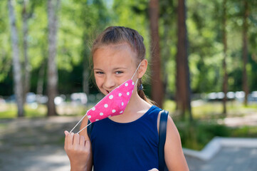 Portrait of a happy caucasian little girl taking off face mask outdoors. Finally, quarantine will...