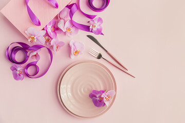 Flower and table settings overhead composition on light pink background. Pink ceramic plates, cutlery, pink gift bag with purple ribbons and pink orchid flowers. Holiday modern table decoration
