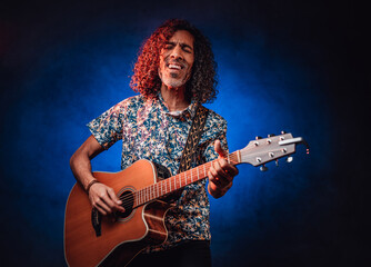 Obraz premium Hispanic musician emotionally singing and playing guitar on a dark lluminated by blue and red light. Concept of music, hobby, festival