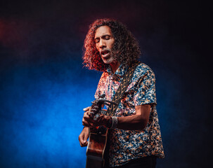 Middle aged hispanic man musician in a hawaiian shirt playing guitar on a dark illuminated by blue and red light. Concept of music, hobby, festival