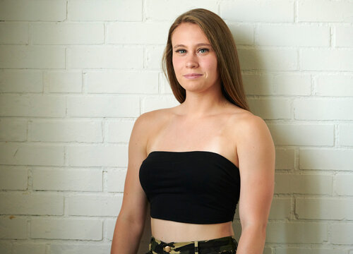 Beautiful young woman in camouflage pants and strapless top poses near window and white brick wall