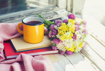 A bouquet of wild flowers, a сup and the books on the vintage windowsill.