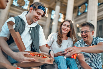 A group of students sit on the steps outside the campus and eat pizza and soda. A group of friends are relaxing and chatting.