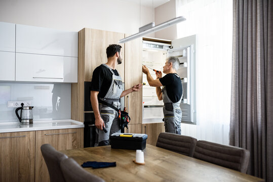 Adding care to your technologies. Aged repairman in uniform fixing refrigerator in the kitchen, while his colleague helping him, bringing screwdriver. Repair service concept