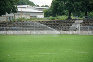 old football stadium with a tribune with decayed wooden seats or banks in a provincial town in Eastern Europe. The lawn, playing field is being irrigated to keep the grass in good condition.