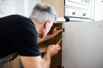 Close up shot of aged repairman in uniform working, fixing kitchen cabinet using screwdriver. Repair service concept