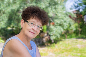 An elderly woman in glasses sits in nature and looks at the camera. The concept of old age, wisdom and joy.