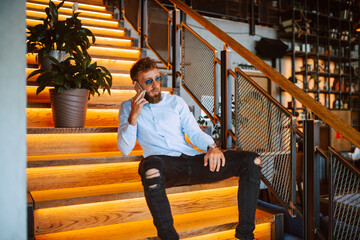 Fototapeta na wymiar A young caucasian man with sunglasses sitting on the steps in a cafe is talking on the phone. Handsome businessman in a restaurant