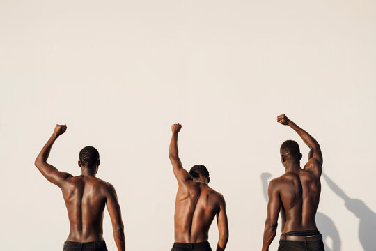 Three men raising fists in protest, standing against white wall, rear view