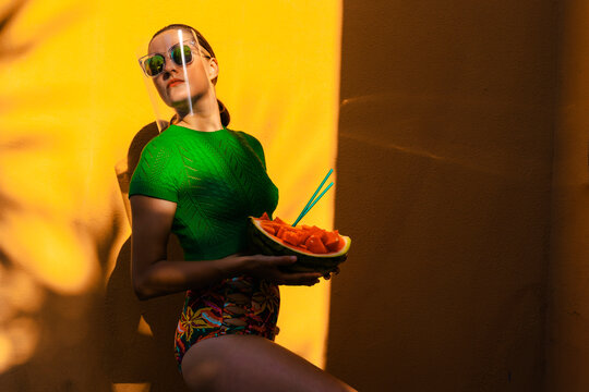 Woman in colorful backyard holding watermelon, wearing sunglasses uner face shield