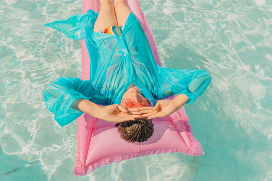 Woman wearing blue rain coat relaxing on pink airbed in swimming pool