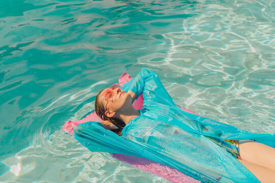 Woman wearing blue rain coat relaxing on pink airbed in swimming pool