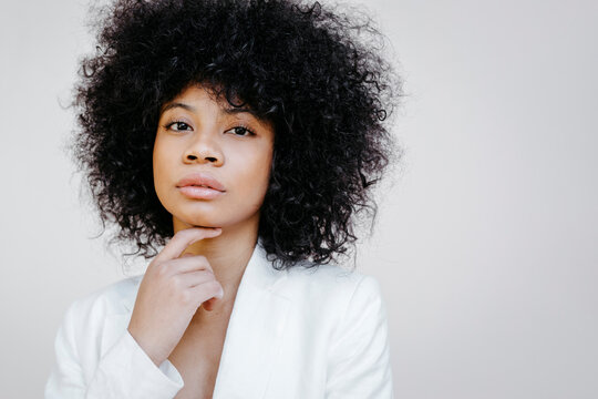 Black womam, wearing white suit, standing in front of white wall, looking pensive