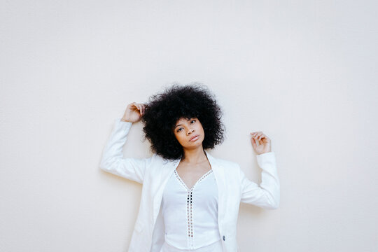 Black womam, wearing white suit, leaning on white wall