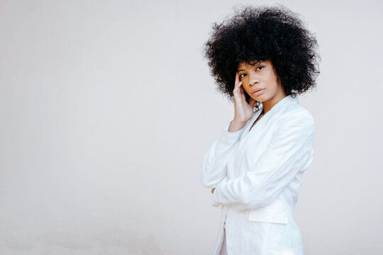 Black womam, wearing white suit, standing in front of white wall, looking pensive